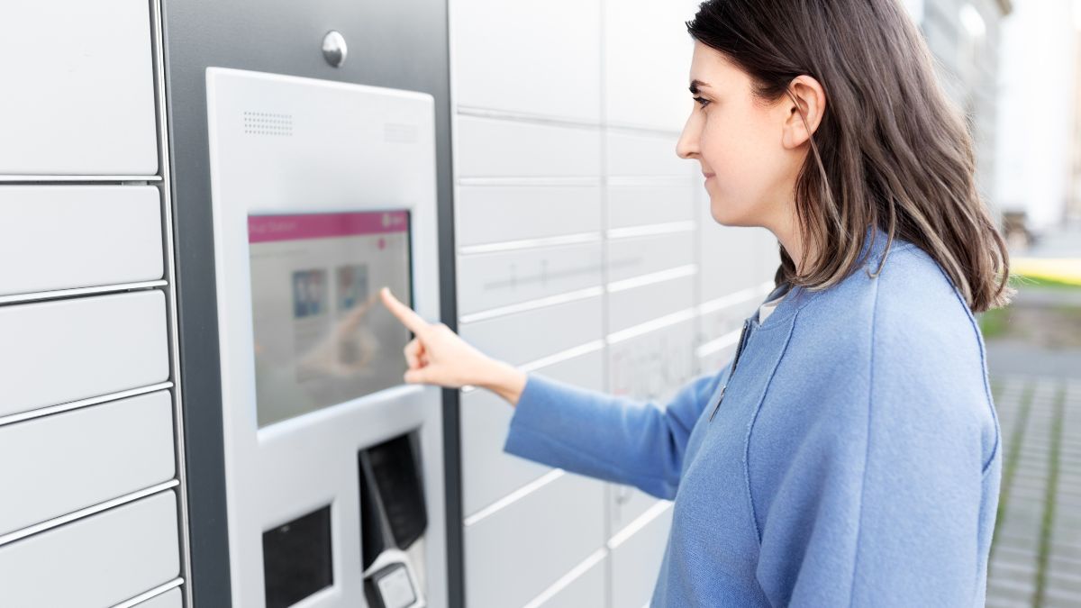 Egy nő az Alzabox automatizált csomagátvételi rendszerénél áll, érintőképernyőt használva.