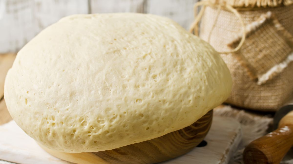 Friss, megkelt tészta egy fából készült tálban, háttérben konyhai eszközök.