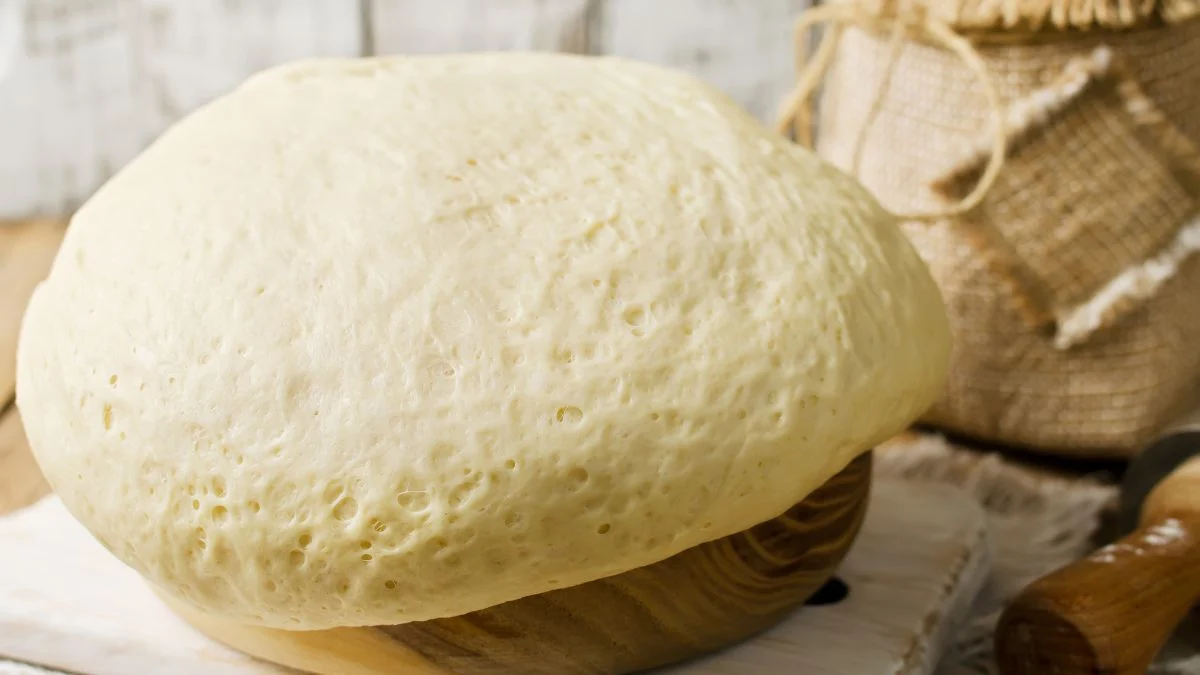 Friss, megkelt tészta egy fából készült tálban, háttérben konyhai eszközök.