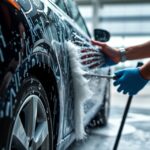 Kézi autómosás művelet, víz és hab a fekete autó mellett.